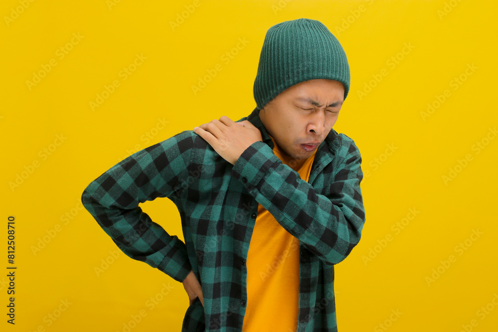 Asian man, dressed in a beanie hat and casual outfit, is seen massaging his shoulder, clearly experiencing backache and shoulder pain due to overwork, isolated on a yellow background. Asian man, dressed in a beanie hat and casual outfit, is seen massaging his shoulder, clearly experiencing backache and shoulder pain due to overwork, isolated on a yellow background.