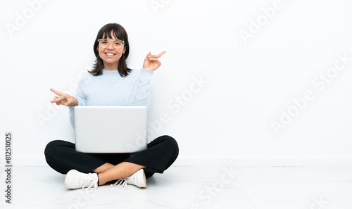 Young mixed race woman sitting on the floor with laptop isolated on white background pointing finger to the laterals and happy © luismolinero