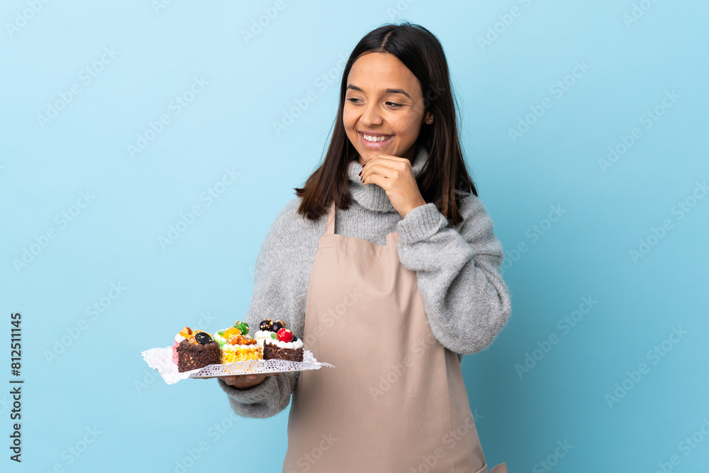 Pastry chef holding a big cake over isolated blue background looking to the side and smiling. Pastry chef holding a big cake over isolated blue background looking to the side and smiling.