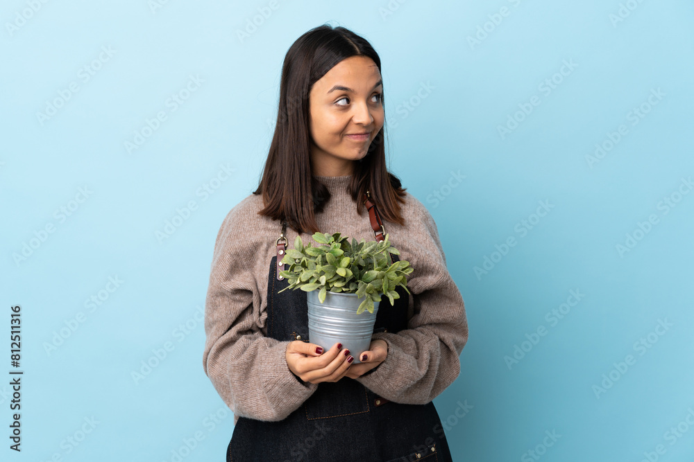 Young brunette mixed race woman holding a plant over isolated blue background scheming something. Young brunette mixed race woman holding a plant over isolated blue background scheming something.