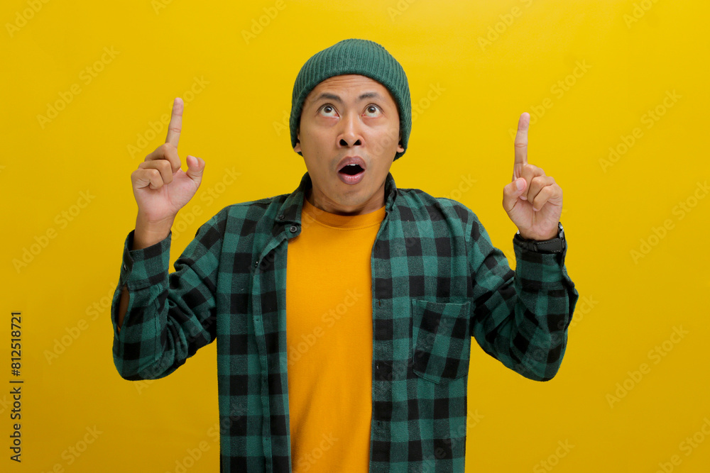 Astonished Asian man, dressed in a beanie hat and casual shirt, points at an empty space, possibly promoting a product or sharing information about an incredible offer, isolated on yellow background. Astonished Asian man, dressed in a beanie hat and casual shirt, points at an empty space, possibly promoting a product or sharing information about an incredible offer, isolated on yellow background.