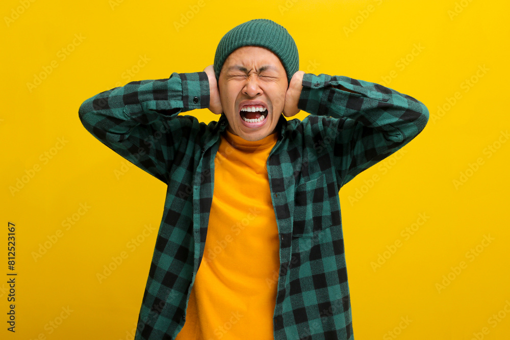 Annoyed young Asian man, wearing a beanie hat and casual shirt, covers his ears and appears to be screaming or shouting, experiencing a painful earache possibly due to otitis exacerbated by loud noise Annoyed young Asian man, wearing a beanie hat and casual shirt, covers his ears and appears to be screaming or shouting, experiencing a painful earache possibly due to otitis exacerbated by loud noise