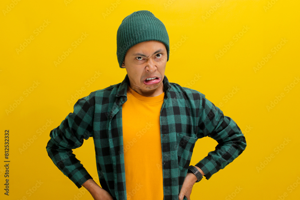 Displeased young Asian man, dressed in a beanie hat and casual shirt, is clearly expressing his disgust at something awful while standing against yellow background. Displeased young Asian man, dressed in a beanie hat and casual shirt, is clearly expressing his disgust at something awful while standing against yellow background.