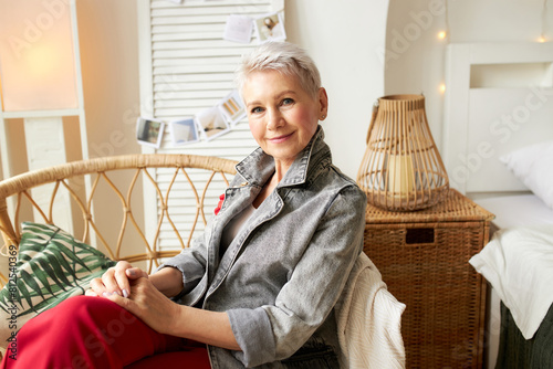 Elegant caucasian senior lady having rest after decorating her country house according to modern trendy interior design, sitting in wicker chair in denim jacket and red pants, putting hands on knees