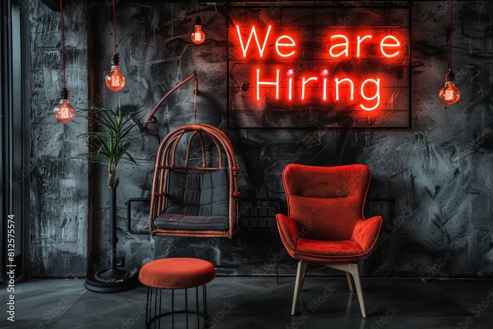 Naklejka premium A vibrant neon sign stating We are hiring stands in front of a red chair, inviting potential candidates to apply for opportunities in a dynamic office setting.