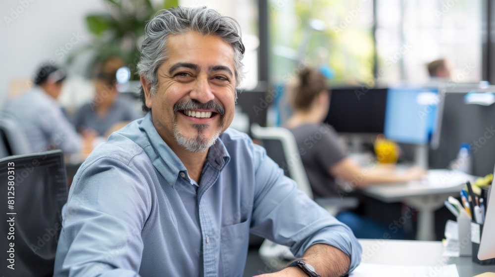 Smiling man with gray hair and beard wearing a blue shirt sitting at a desk in an office environment with colleagues in the background. Smiling man with gray hair and beard wearing a blue shirt sitting at a desk in an office environment with colleagues in the background.