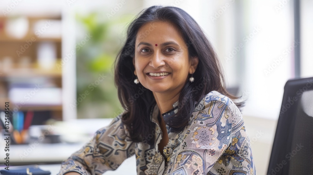 Smiling woman with dark hair wearing a patterned blouse sitting at a desk in an office environment. Smiling woman with dark hair wearing a patterned blouse sitting at a desk in an office environment.
