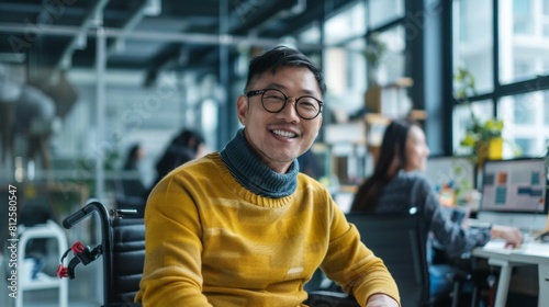 Smiling man in yellow sweater and glasses sitting at desk in modern office with large windows and plants looking at camera.
