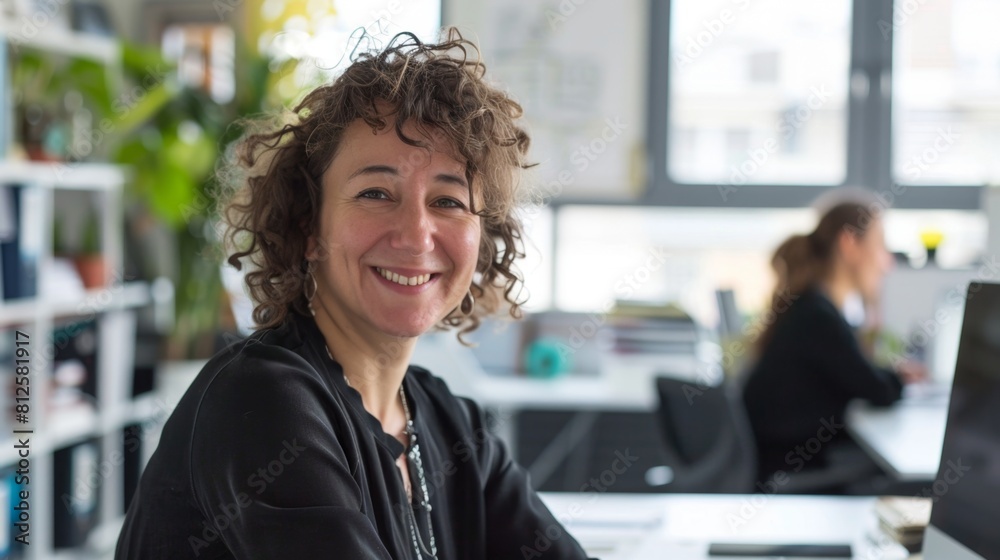 Smiling woman with curly hair wearing black top sitting at desk in modern office with large windows and greenery. Smiling woman with curly hair wearing black top sitting at desk in modern office with large windows and greenery.