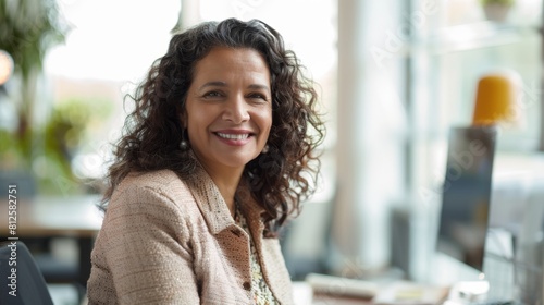 Smiling woman with curly hair wearing a beige jacket sitting at a desk with a blurred background of a modern office.