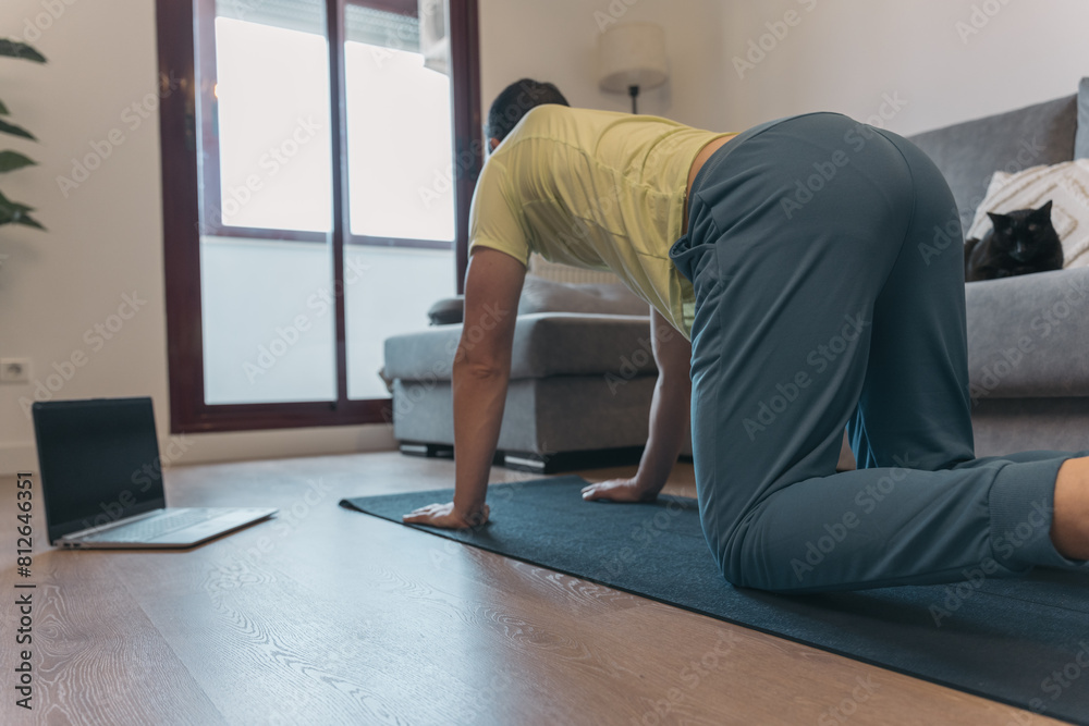 Anonymous Man Stretching on Mat with Laptop for Online Class in Home Setting. Unrecognizable man does yoga stretches on mat with laptop nearby for online class in cozy home environment.