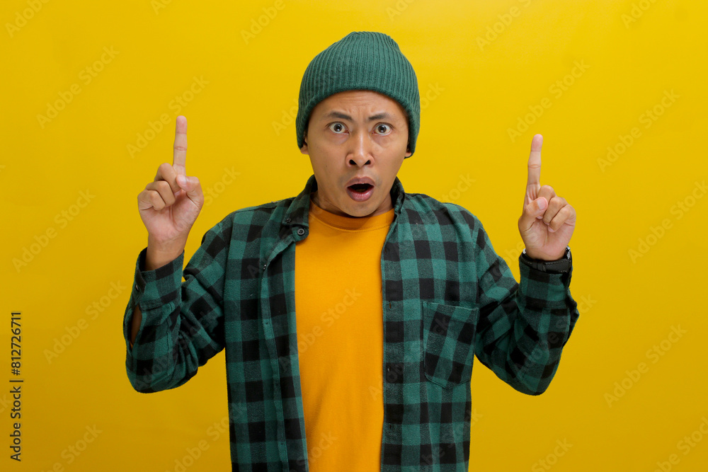 Astonished Asian man, dressed in a beanie hat and casual shirt, points at an empty space, possibly promoting a product or sharing information about an incredible offer, isolated on yellow background.