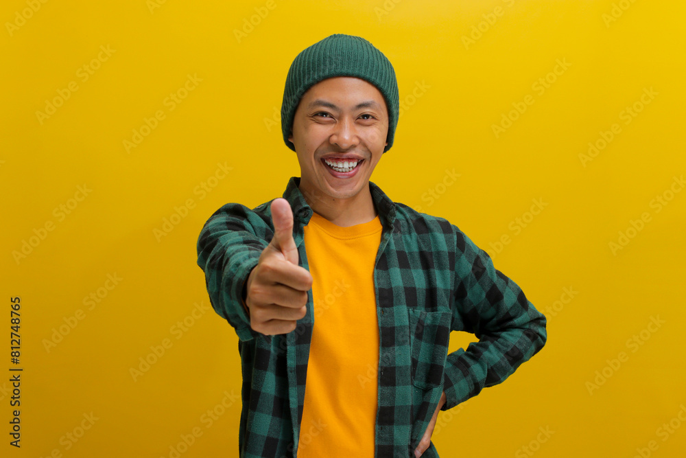 Confident Asian man is expressing approval and recommendation by showing a thumbs-up gesture, indicating his approval or liking for something positive. Standing against yellow background. Confident Asian man is expressing approval and recommendation by showing a thumbs-up gesture, indicating his approval or liking for something positive. Standing against yellow background.