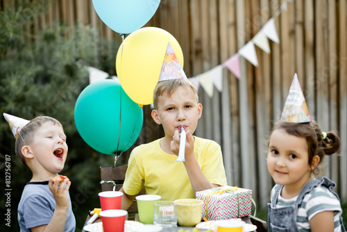 Cute funny nine year old boy celebrating his birthday with family or friends with homemade baked cake in a backyard. Birthday party. Kids wearing party hats and blowing whistles.