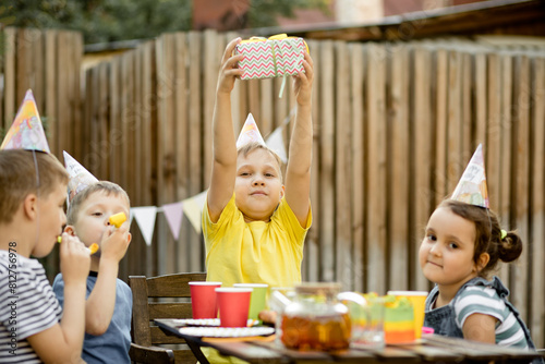 Cute funny nine year old boy celebrating his birthday with family or friends in a backyard. Birthday party. Kid wearing party hat and holding gift box.