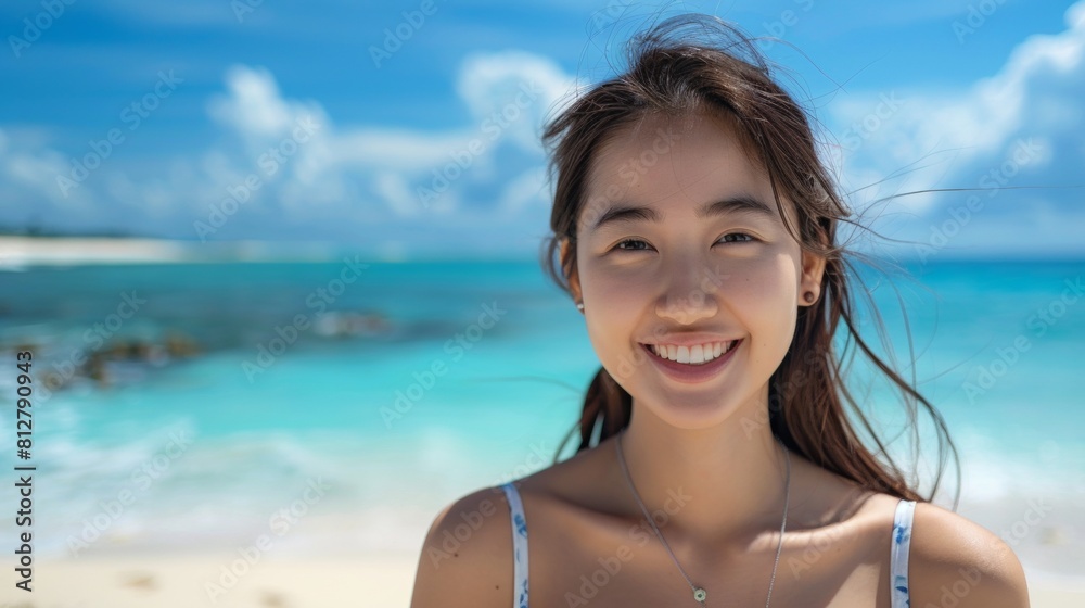 Joyful Asian woman smiling on a sunny beach, with her hair tousled by the breeze, against a backdrop of vibrant turquoise waters.