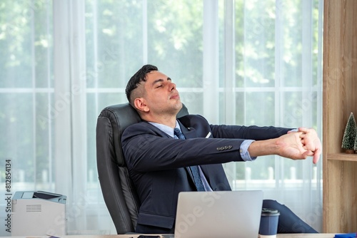 A man in a suit is sitting in an office chair with his eyes closed and his arms outstretched. He is wearing a tie and a suit jacket. He has a laptop on his desk. © BESTIMAGE