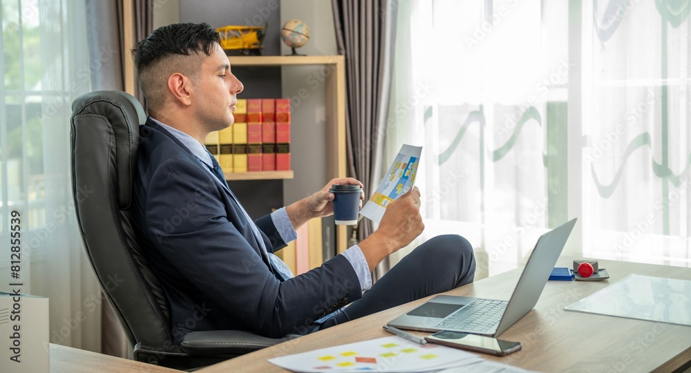A man in a suit is sitting in a chair with his feet on the desk, holding a cup of coffee and looking at a document. A man in a suit is sitting in a chair with his feet on the desk, holding a cup of coffee and looking at a document.