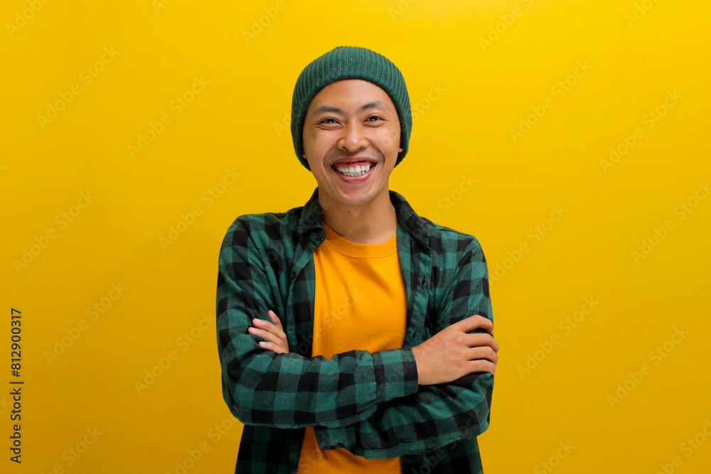 Confident young Asian man, clad in a beanie hat and casual outfit, stands with folded arms, wearing a confident smile as he gazes at the camera while standing against a yellow background Confident young Asian man, clad in a beanie hat and casual outfit, stands with folded arms, wearing a confident smile as he gazes at the camera while standing against a yellow background