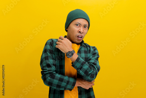 Displeased young Asian man, dressed in a beanie hat and casual shirt, is clearly expressing his disgust at something awful while standing against yellow background. photo