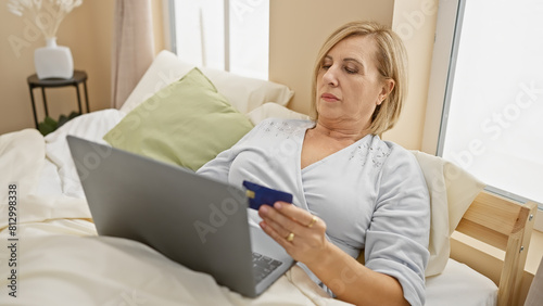 A middle-aged woman reclines in bed with a laptop and credit card  encapsulating leisure  technology  and shopping in a home setting.