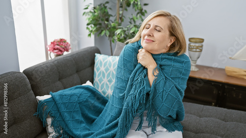 Blonde woman relaxing on a couch at home draped in a teal throw, eyes closed with a contented smile.