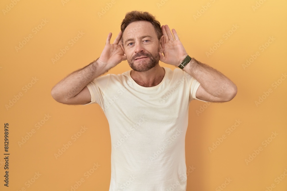 Middle age man with beard standing over yellow background trying to hear both hands on ear gesture, curious for gossip. hearing problem, deaf Middle age man with beard standing over yellow background trying to hear both hands on ear gesture, curious for gossip. hearing problem, deaf