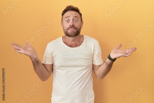 Middle age man with beard standing over yellow background clueless and confused expression with arms and hands raised. doubt concept.