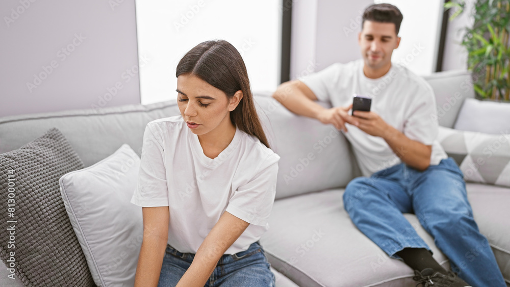 A woman looks upset while a man uses his phone, indicating relationship issues in a modern living room. A woman looks upset while a man uses his phone, indicating relationship issues in a modern living room.