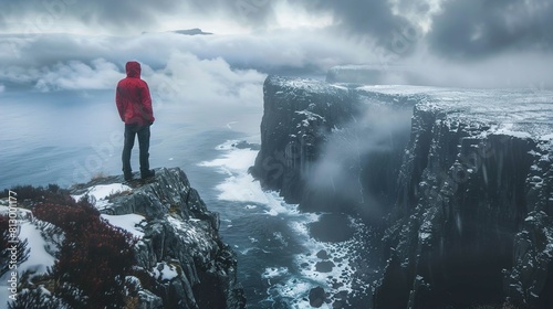 A person in a red jacket and dark pants stands on a rugged cliff edge, facing a dramatic seascape. The scene is enveloped in a mix of low-hanging clouds and mist, with the sea stretching out to the ho