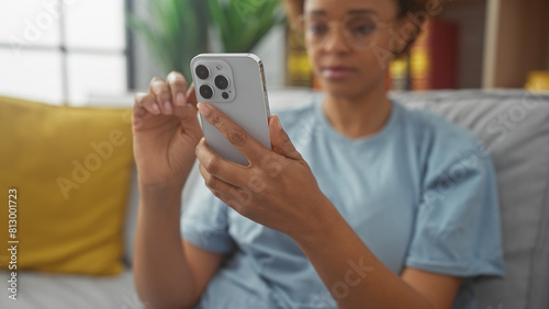 African american woman uses smartphone, sitting indoors on a couch in a well-lit living room, exhibiting casual, modern lifestyle.