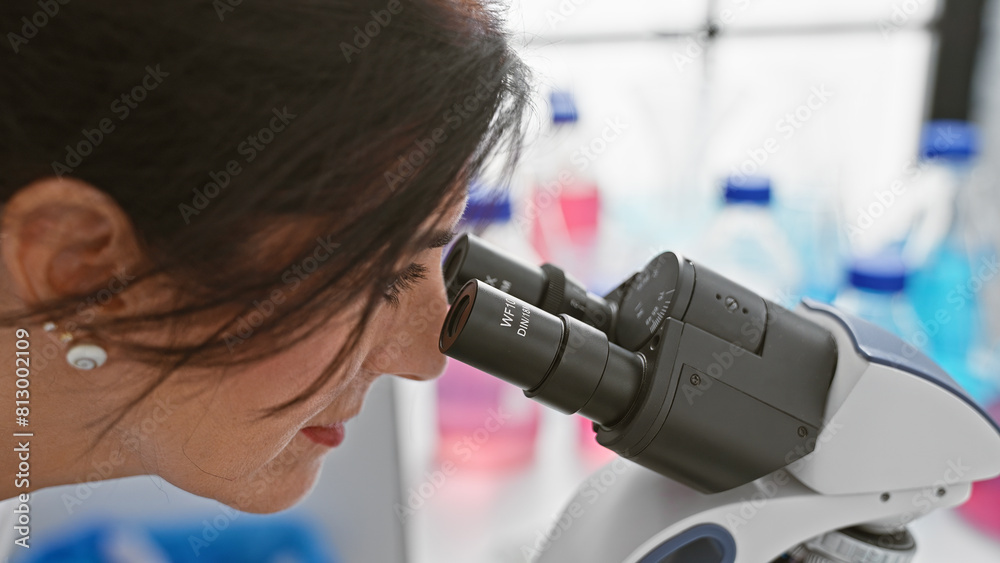 A brunette woman examines samples through a microscope in a laboratory setting with blurred background.