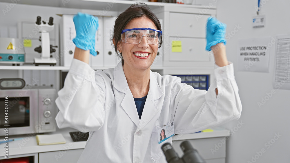 A smiling hispanic woman donning lab coat and safety goggles poses triumphantly in a well-equipped laboratory setting. A smiling hispanic woman donning lab coat and safety goggles poses triumphantly in a well-equipped laboratory setting.