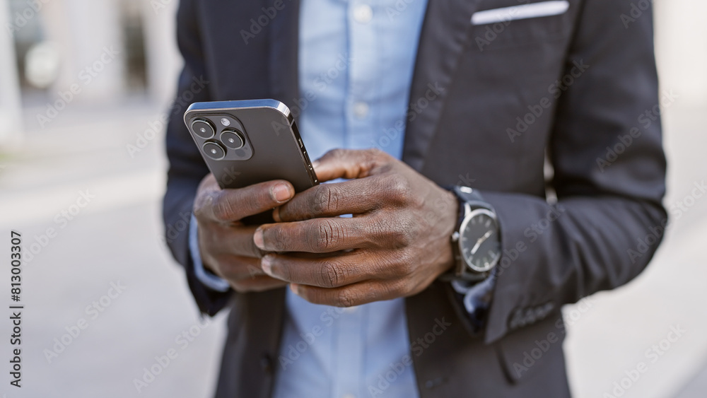 An african american man in a stylish suit uses a smartphone on a sunny city street. An african american man in a stylish suit uses a smartphone on a sunny city street.