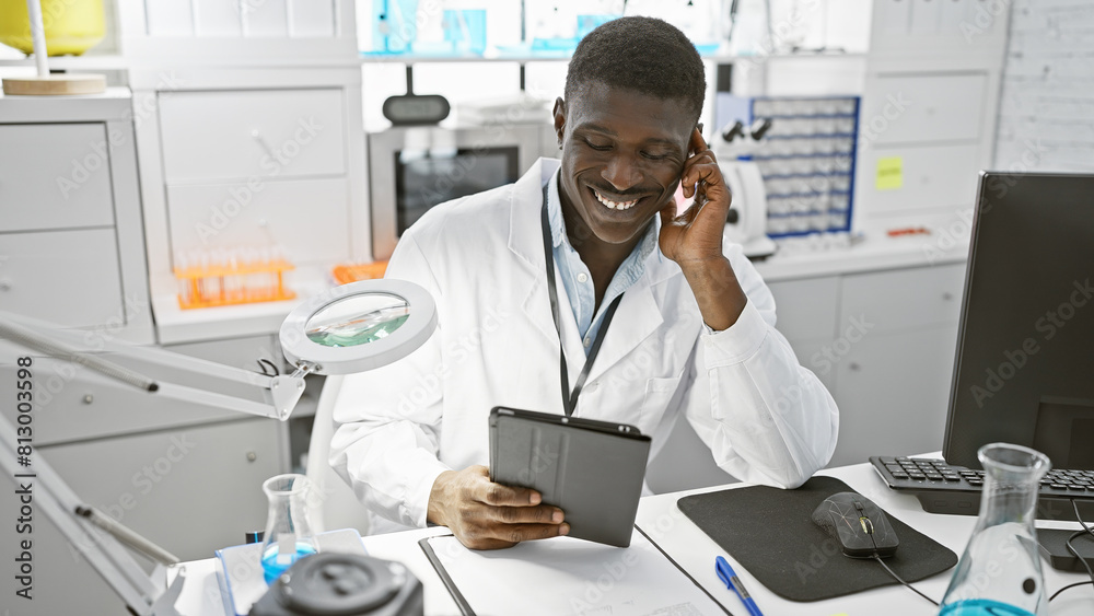 A smiling african man in a lab coat using a tablet in a modern laboratory takes a phone call.