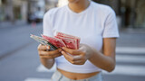 A young asian woman counts chinese yuan banknotes on a bustling city street.
