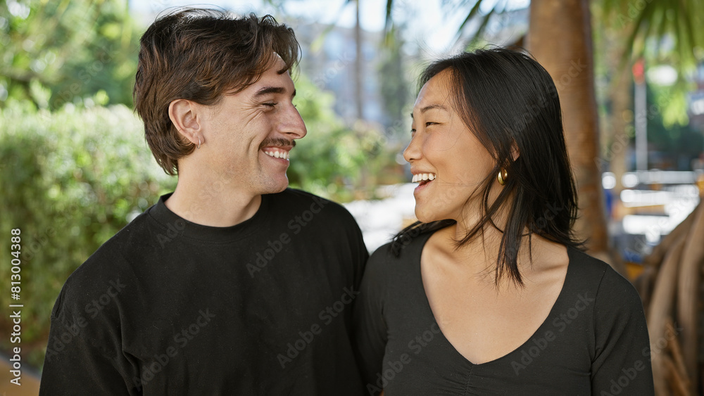 Interracial couple smiling together in a sunny park, portraying love and happiness with a man and woman outdoors. Interracial couple smiling together in a sunny park, portraying love and happiness with a man and woman outdoors.