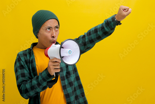 Asian man activist in a beanie hat shouts and yells into a megaphone, demanding change and protesting for human rights or justice with a focus on freedom of speech. Isolated on a yellow background photo