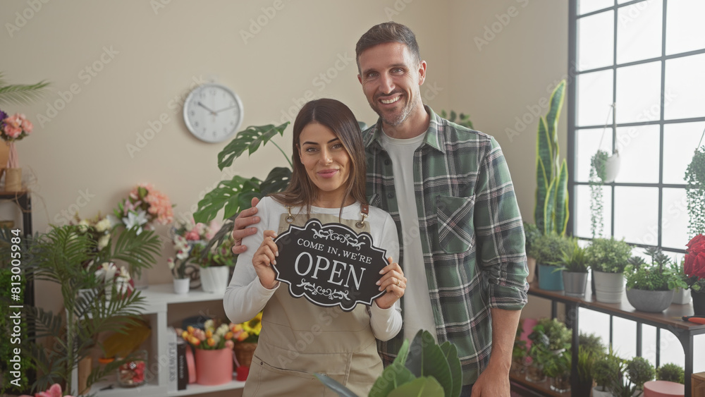 Fototapeta premium Smiling man and woman holding an 'open' sign in a vibrant indoor flower shop filled with various plants.