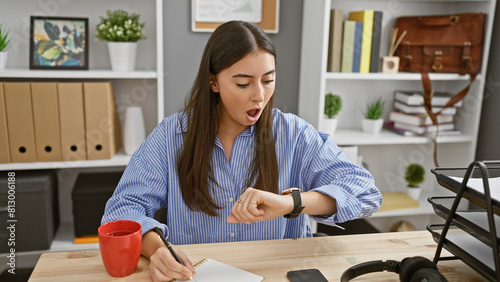 Fotografi A surprised hispanic woman checks the time in a modern office environment, illustrating punctuality and deadline awareness