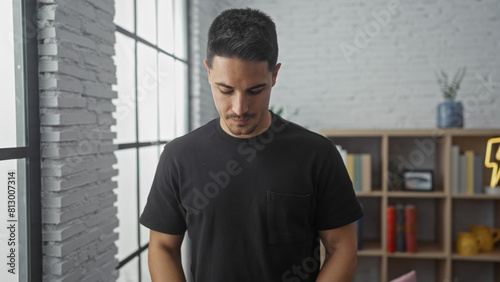 Hispanic man with beard in black shirt stands thoughtfully in a modern living room.