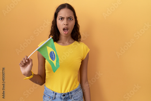 Young hispanic woman holding brazil flag in shock face  looking skeptical and sarcastic  surprised with open mouth