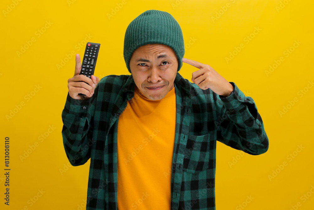 A discontented Asian man, dressed in a beanie hat and casual shirt, covers his ears in response to loud noises while holding a TV remote, isolated on a yellow background A discontented Asian man, dressed in a beanie hat and casual shirt, covers his ears in response to loud noises while holding a TV remote, isolated on a yellow background