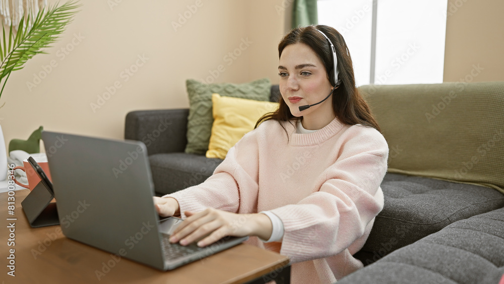Young caucasian woman wearing a headset works on laptop in cozy living room interior. Young caucasian woman wearing a headset works on laptop in cozy living room interior.