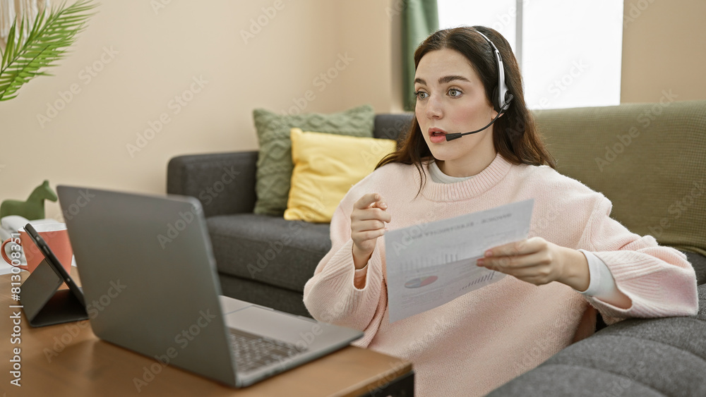 A young caucasian woman is working from home, conducting a video call while holding a document, with a laptop on her desk in an apartment living room. A young caucasian woman is working from home, conducting a video call while holding a document, with a laptop on her desk in an apartment living room.