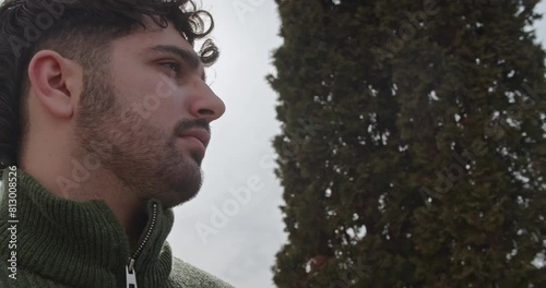 A young man in a green sweater thoughtfully removes his glasses in a peaceful outdoor environment, suggesting a moment of reflection or a pause in his busy day.
