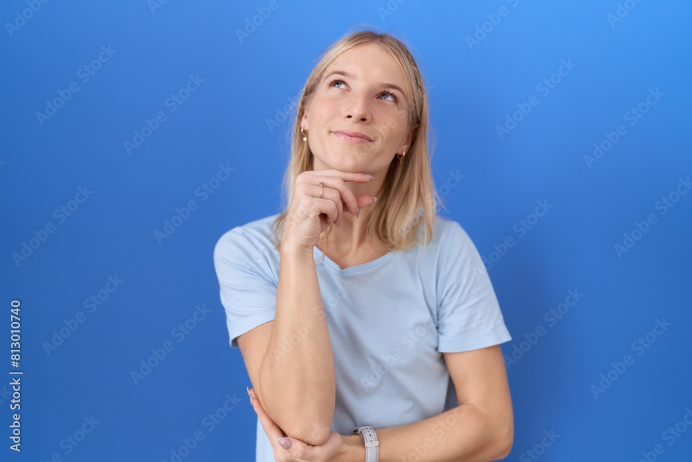 Young caucasian woman wearing casual blue t shirt with hand on chin thinking about question, pensive expression. smiling and thoughtful face. doubt concept. Young caucasian woman wearing casual blue t shirt with hand on chin thinking about question, pensive expression. smiling and thoughtful face. doubt concept.