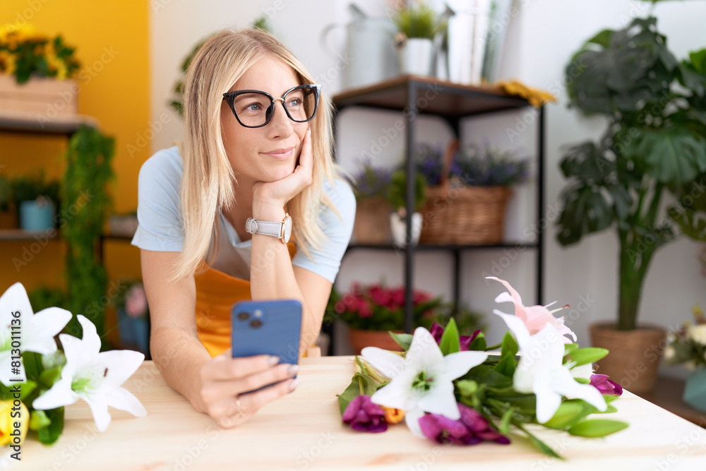 Young blonde woman florist using smartphone leaning on table at flower shop