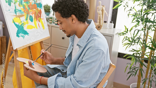 Young adult man with curly hair painting on canvas in art studio, showcasing creativity and concentration. photo