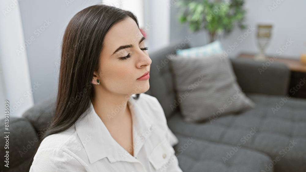 Portrait of a serene hispanic woman with closed eyes in a white shirt, indoors at home with a couch and cushion in the background Portrait of a serene hispanic woman with closed eyes in a white shirt, indoors at home with a couch and cushion in the background